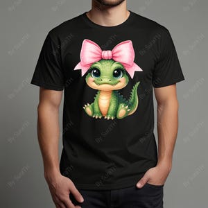 May include: Black t-shirt featuring a cute cartoon green alligator with a pink bow on its head. The alligator has large eyes and is smiling. The shirt is made of a soft material.