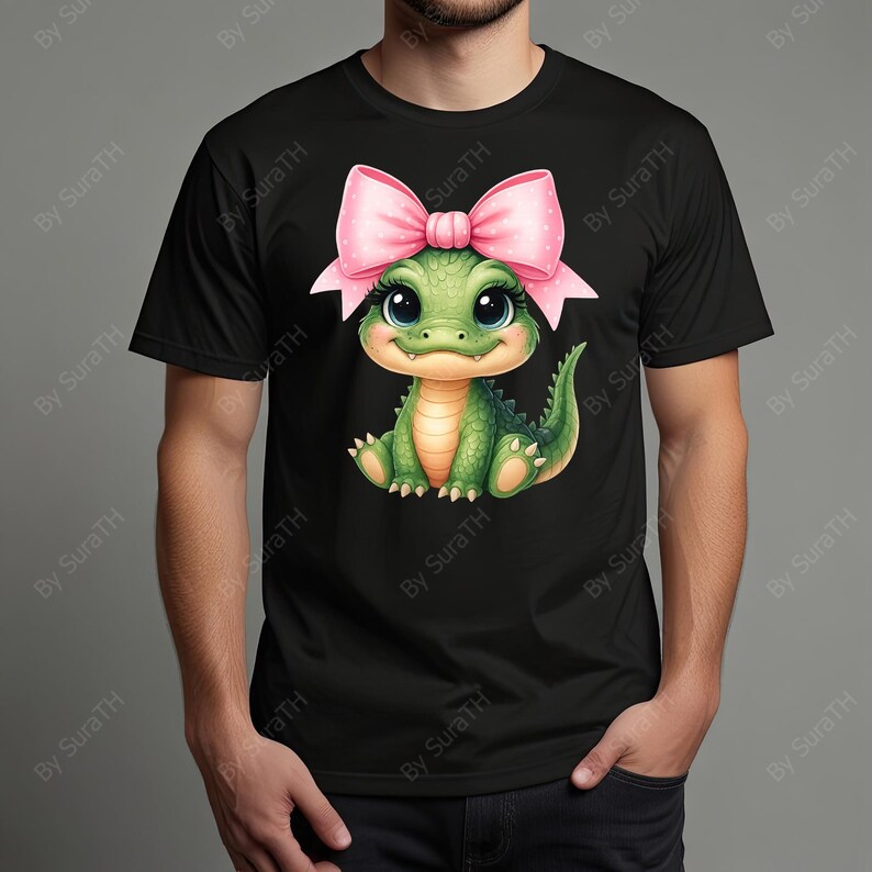 May include: Black t-shirt featuring a cute cartoon green alligator with a pink bow on its head. The alligator has large eyes and is smiling. The shirt is made of a soft material.