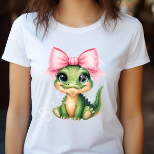 May include: White t-shirt featuring a cartoon alligator with a pink bow. The alligator is green with a light orange belly and large, expressive eyes. The bow is pink with white polka dots. The design is centered on the shirt.
