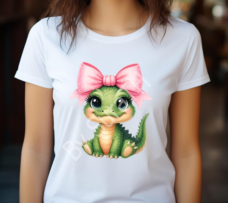 May include: White t-shirt featuring a cartoon alligator with a pink bow. The alligator is green with a light orange belly and large, expressive eyes. The bow is pink with white polka dots. The design is centered on the shirt.
