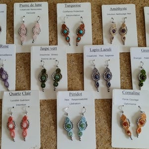 May include: A collection of 12 pairs of handmade earrings featuring various gemstone colors and designs. Each pair is displayed on a white card with the name of the gemstone and its associated meaning in French.