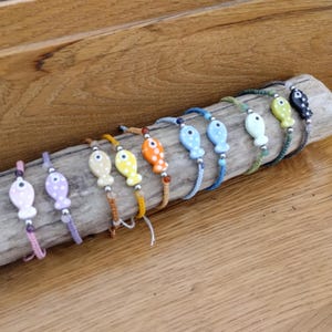 Peut inclure: Une collection de bracelets faits à la main en forme de poisson, de différentes couleurs, dont rose, violet, jaune, orange et bleu. Chaque bracelet comporte un charm en forme de poisson à pois et est monté sur un cordon. Les bracelets sont exposés sur un morceau de bois flotté.