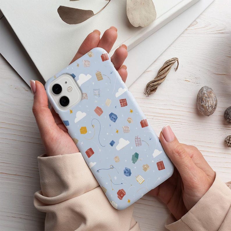 May include: A light blue phone case with a pattern of colorful gift boxes, white clouds, and yellow stars. The case is held in a person's hands. The phone case is designed for a smartphone.