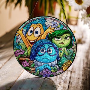May include: Round stained glass panel featuring characters from the animated movie "Inside Out." The panel depicts Joy, Sadness, and Disgust surrounded by flowers in a garden setting. The panel is suspended by a chain.