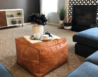 May include: A square, tan leather ottoman sits in the center of a room. On top of the ottoman is a white faux fur throw, a round wooden tray, a white textured vase with dark flowers, a small candle, and a stack of white coasters.