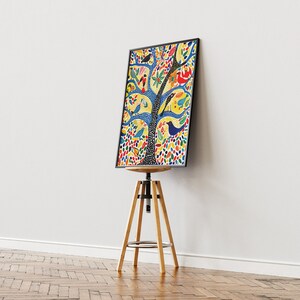 Op de afbeelding: Een kleurrijke ingelijste print van een boom met vogels en bloemen op een gele achtergrond. De print wordt getoond op een houten kruk.