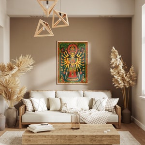 May include: A living room interior design with a white couch, a wooden coffee table, and a framed print of a colorful Hindu deity with multiple arms. The print is set against a brown wall with a light brown ceiling. There are two potted plants, one with dried palm leaves and the other with dried pampas grass.