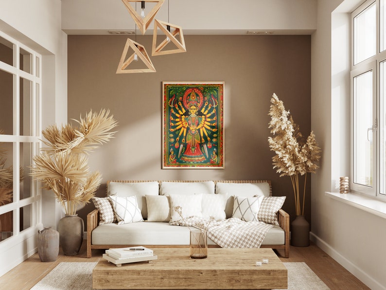 May include: A living room interior design with a white couch, a wooden coffee table, and a framed print of a colorful Hindu deity with multiple arms. The print is set against a brown wall with a light brown ceiling. There are two potted plants, one with dried palm leaves and the other with dried pampas grass.