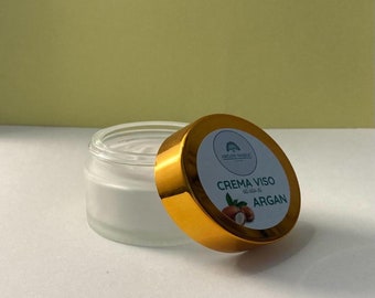 May include: A gold jar with a white label that reads "Crema Viso al olio di Argan" and a green logo of a tree. The jar is open and the lid is off. The jar is on a white surface.