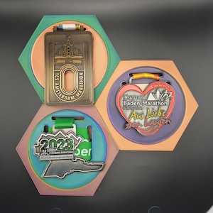 Könnte beinhalten: Drei sechseckige Medaillenhalter mit drei verschiedenen Medaillen. Die oberste Medaille ist aus Bronze und trägt den Text "TCS-AMSTERDAM MARATHON" und "2023". Die mittlere Medaille ist lila und trägt ein Herz mit dem Text "26 ATTUVIA Baden-Marathon Karlsruhe Aus Liebe zum Laufen" und "Version 27". Die unterste Medaille ist silber und trägt den Text "2023 FREIBURG MARATHON #meinfreiburgmarathon".