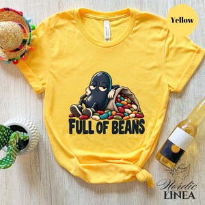 Puede incluir: Una camiseta amarilla de manga corta con un gráfico de un personaje de frijol negro apoyado contra un saco de arpillera lleno de frijoles de colores. Debajo del gráfico, el texto dice FULL OF BEANS en letras mayúsculas negras.