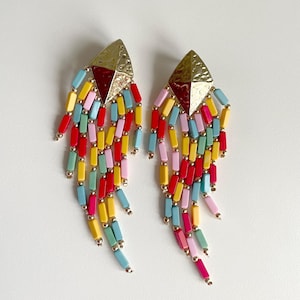Op de afbeelding: Een paar kleurrijke oorhangers. Elke oorbel heeft een goudkleurige, diamantvormige bovenkant met een textuuroppervlak. Daaronder vallen strengen kleurrijke kralen in rood, geel, roze, blauw en groen naar beneden, wat een levendig ontwerp creëert.