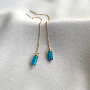 Op de afbeelding: Een paar goudkleurige oorhangers met turquoise rechthoekige kralen. De oorbellen hangen aan fijne kettingen, met een kleine gouden kraal aan de onderkant van elke turquoise kraal. De oorbellen zijn afgebeeld tegen een witte achtergrond.