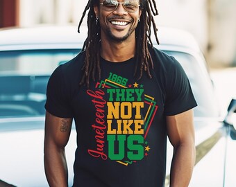 Può includere: T-shirt nera con la scritta "Juneteenth" in rosso sul lato sinistro. La maglietta presenta la scritta "1865 THEY NOT LIKE US" in forme geometriche gialle, verdi e rosse. La persona indossa occhiali da sole e un orologio.