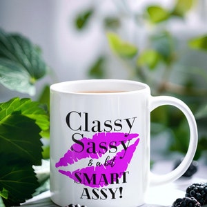 以下が含まれることがあります： 白地に黒とピンクのデザインが施されたセラミック製のマグカップ。マグカップには「Classy Sassy & a bit SMART ASSY!」という文字が印刷されています。デザインにはピンクの唇のプリントが含まれています。
