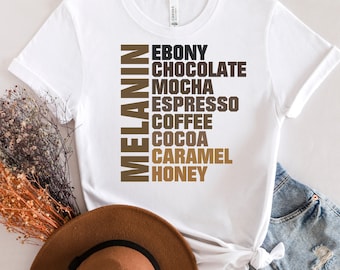 May include: White t-shirt with the word "MELANIN" written vertically in brown letters. The words "EBONY", "CHOCOLATE", "MOCHA", "ESPRESSO", "COFFEE", "COCOA", "CARAMEL", and "HONEY" are written vertically in brown letters below the word "MELANIN".