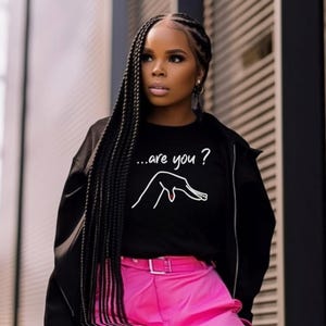 Puede incluir: Sudadera negra de cuello redondo con el texto "...are you?" y un gráfico de gesto de la mano. La sudadera se usa con una chaqueta negra y pantalones rosas. La imagen está tomada al aire libre.