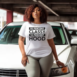 Puede incluir: Una camiseta blanca con texto negro que dice "90 Percent Classy 10 Percent Hood". La persona que lleva la camiseta está de pie frente a un coche blanco.