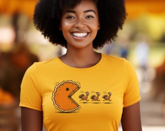 Puede incluir: Una mujer lleva una camiseta amarilla con un diseño inspirado en Pac-Man. El gráfico presenta una porción de pastel persiguiendo a tres pavos. La mujer sonríe, con un fondo borroso naranja y verde.