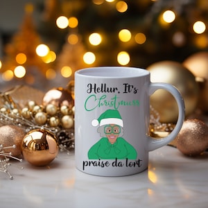 Könnte beinhalten: Weiße Keramiktasse mit einer grünen und schwarzen Cartoon-Illustration einer Frau, die einen grünen Weihnachtsmann-Hut und eine Brille trägt. Der Text "Hellur. It's Christmas praise da lord" ist auf die Tasse gedruckt.