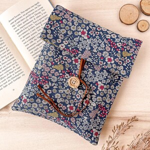 May include: A blue fabric book cover with a floral pattern. The cover has a brown leather strap and a wooden button closure.