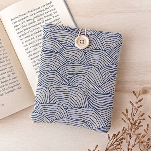 Può includere: Una copertina per libro in tessuto beige con un motivo a onde blu e bianco. La copertina ha una chiusura a bottone e un cinturino bianco.