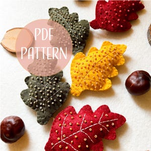 Può includere: Modello PDF per ornamenti a foglia di feltro. Le foglie sono in tonalità di rosso, giallo e verde e sono decorate con piccole perline. Il testo "PDF PATTERN" è scritto in un cerchio rosa.