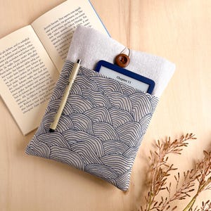 Op de afbeelding: Een stoffen e-reader hoes met een blauw en wit golfpatroon, met een e-reader met "Chapter 11". Een pen en een boek liggen naast de hoes. De hoes is vastgezet met een houten knoop.