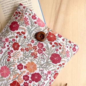 May include: A floral book sleeve with a white background and a pattern of red, pink, and orange flowers. The sleeve has a wooden button closure and is partially covering a book.