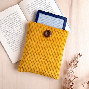 Può includere: Custodia per e-reader in velluto a coste giallo senape con chiusura a bottone marrone. La custodia contiene un e-reader ed è posizionata sopra un libro aperto. Un rametto di fiori secchi è sullo sfondo.