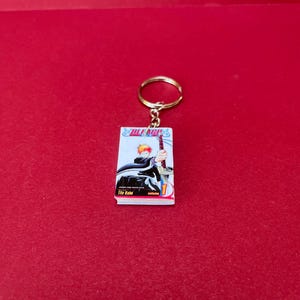 May include: A rectangular keychain displaying the cover of a manga volume. The illustration shows a character with orange hair wielding a sword. The keychain is attached to a gold-coloured ring.