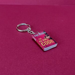 May include: A silver keyring with a miniature book charm. The book is a replica of "The Da Vinci Code" by Dan Brown, with a red and white cover and a man's face. The keyring is on a deep red background.