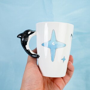 Puede incluir: Taza de cer&aacute;mica blanca con un asa de orca en blanco y negro. La taza tiene un dise&ntilde;o de acuarela azul de una estrella con una luna creciente en su interior. Hay dos estrellas blancas m&aacute;s peque&ntilde;as en la taza.