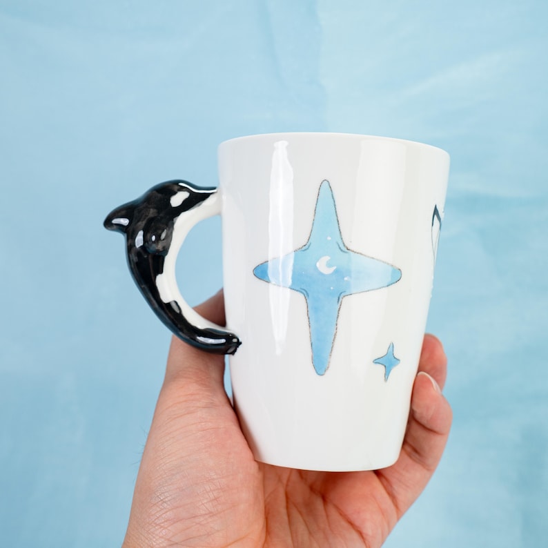 Puede incluir: Taza de cer&aacute;mica blanca con un asa de orca en blanco y negro. La taza tiene un dise&ntilde;o de acuarela azul de una estrella con una luna creciente en su interior. Hay dos estrellas blancas m&aacute;s peque&ntilde;as en la taza.