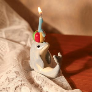 Könnte beinhalten: Eine Keramik-Delfinfigur mit einem bunten Ball auf dem Kopf, der eine brennende blaue Geburtstagskerze mit der Nummer eins hält. Der Delfin ist hellblau mit weißem Bauch, auf weißem Spitzentuch und Holzuntergrund.