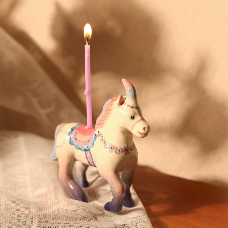 Op de afbeelding: Een keramieken carrousel paardenbeeldje met een roze kaars die op zijn rug brandt. Het paard is geschilderd in pastelkleuren met blauwe, roze en cr&egrave;mekleurige accenten. De kaars brandt met een heldergele vlam.