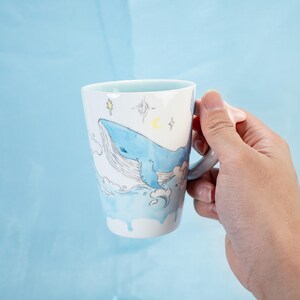 Puede incluir: Una taza de cer&aacute;mica blanca con una ilustraci&oacute;n de una ballena azul y un dise&ntilde;o similar a la acuarela. La ballena nada en un oc&eacute;ano azul con nubes blancas y estrellas. La taza tiene un asa y est&aacute; siendo sostenida por una mano.