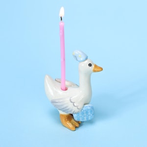 May include: A ceramic goose birthday candle holder with a lit pink candle. The goose is white with a blue hat and a blue basket. The candle is pink and has a flame at the top. The background is light blue.