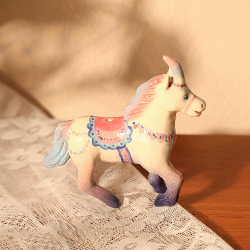 Op de afbeelding: Een keramieken paardenbeeldje in een wandelhouding. Het paard is geschilderd in pastelkleuren, met een roze manen en staart, en een blauw en roze zadel. Het beeldje heeft een grillig, decoratief ontwerp.