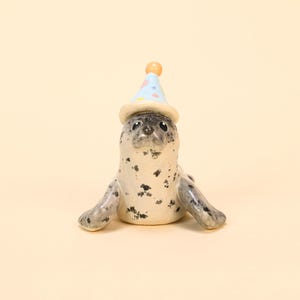 May include: A ceramic seal figurine with a light blue party hat adorned with colorful polka dots and a small orange ball on top. The seal has black spots and outstretched flippers.