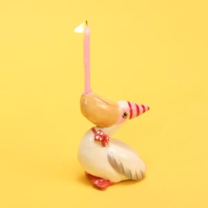 以下が含まれることがあります： 点灯したピンク色のろうそくが付いた、気まぐれなセラミック製のペリカンの誕生日キャンドルホルダー。ペリカンは赤と白のストライプのくちばし、赤い蝶ネクタイ、赤い足を持っています。背景は無地の黄色です。