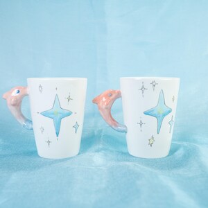 Puede incluir: Dos tazas de cer&aacute;mica blanca con un asa en forma de delf&iacute;n. Las tazas tienen una estrella azul con una luna creciente en su interior y un fondo blanco con detalles de purpurina dorada.