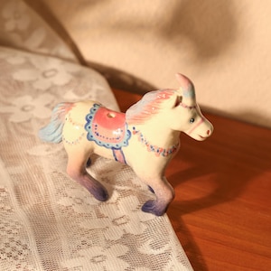 Op de afbeelding: Een keramieken paardenbeeldje met een cr&egrave;mekleurig lichaam, roze en blauwe manen en staart, en een roze zadel. Het paard heeft blauwe en roze accenten en staat in een loopstand. Het beeldje staat op een houten oppervlak.