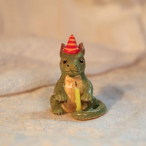K&ouml;nnte beinhalten: Keramikfigur eines gr&uuml;nen Drachen mit roter und orangefarbener Partyh&uuml;tchen. Der Drache hat eine gelbe Kerze vor sich. Die Figur ist detailliert mit einem l&auml;chelnden Gesicht und kleinen H&ouml;rnern. Das Gesamtthema ist eine Geburtstagsfeier.
