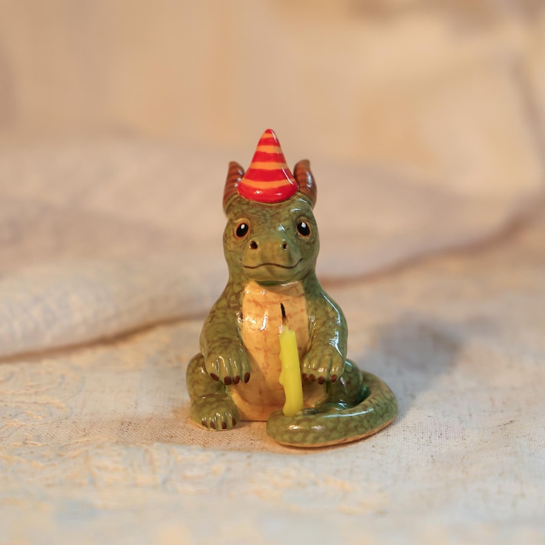 K&ouml;nnte beinhalten: Keramikfigur eines gr&uuml;nen Drachen mit roter und orangefarbener Partyh&uuml;tchen. Der Drache hat eine gelbe Kerze vor sich. Die Figur ist detailliert mit einem l&auml;chelnden Gesicht und kleinen H&ouml;rnern. Das Gesamtthema ist eine Geburtstagsfeier.