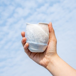 Puede incluir: Una taza de cerámica blanca con un diseño texturizado y en espiral. La taza se sostiene en una mano contra un fondo de cielo azul.