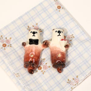 Può includere: Due statuette di lontra in ceramica. Una lontra indossa un papillon nero, l'altra tiene in mano un piccolo bouquet. Le lontre hanno teste bianche e corpi marroni con effetto sfumato. Sono su un fazzoletto a quadri blu e bianco.