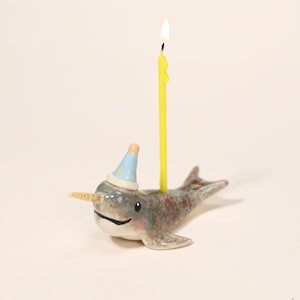 May include: A ceramic narwhal birthday candle holder with a yellow candle. The narwhal is gray, brown, and white with a party hat. The candle is lit with a yellow flame.