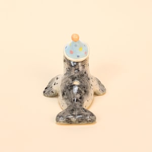 May include: A ceramic seal figurine with a party hat. The seal is gray with black spots and has a light blue party hat with colorful polka dots and a small orange pom-pom on top. The figurine is on a cream-colored background.