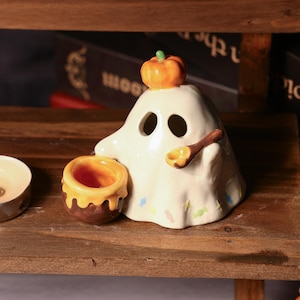 May include: A ceramic Halloween ghost figurine with a small orange pumpkin on its head. The ghost has two eye holes and a spoon-shaped detail. A small bowl with orange glaze sits beside it. The figurine is on a wooden surface.